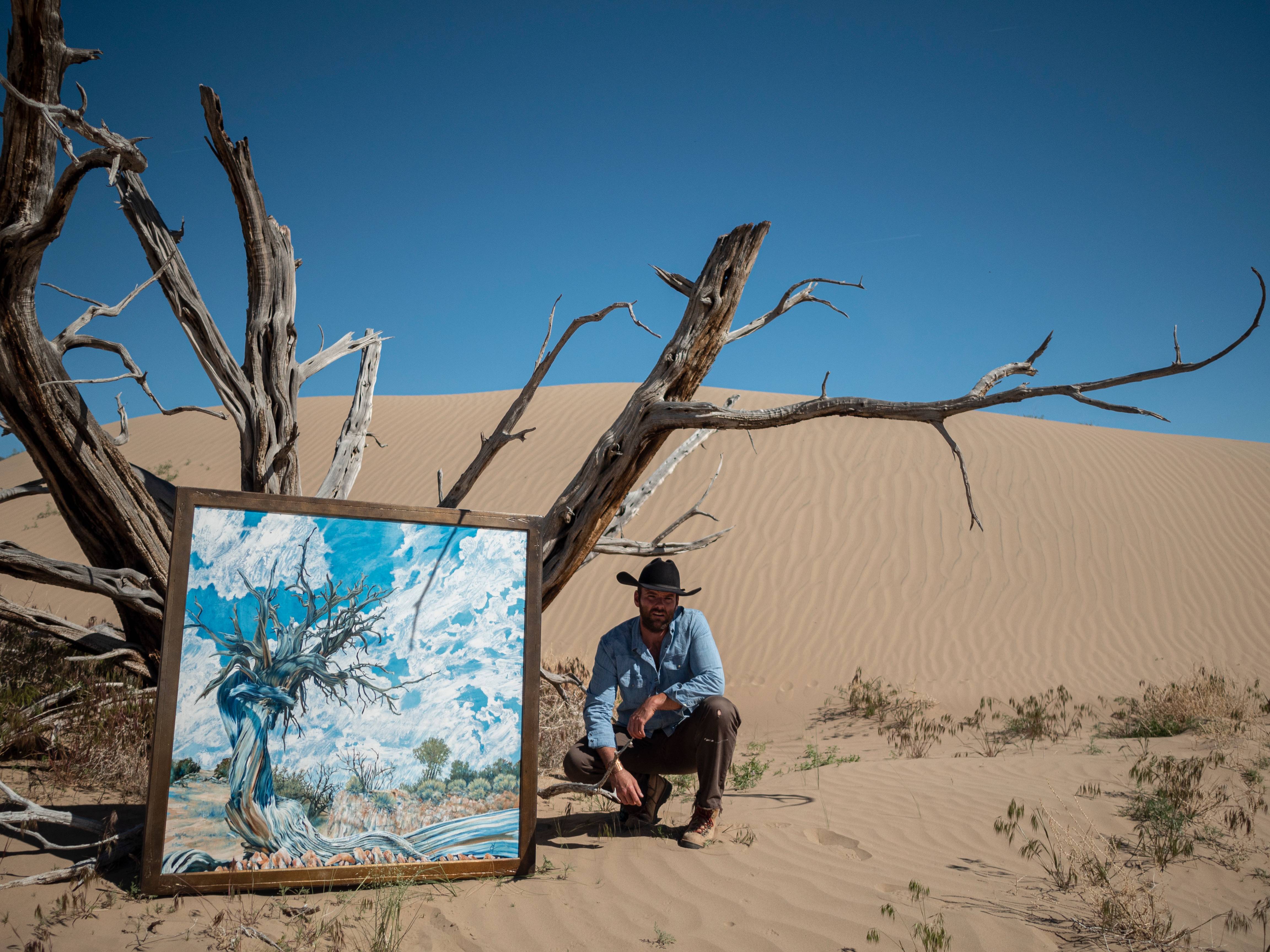 Cody Chamberlain crouching beside the framed Rockwell Ranch Juniper painting and a dead juniper on sand dunes.